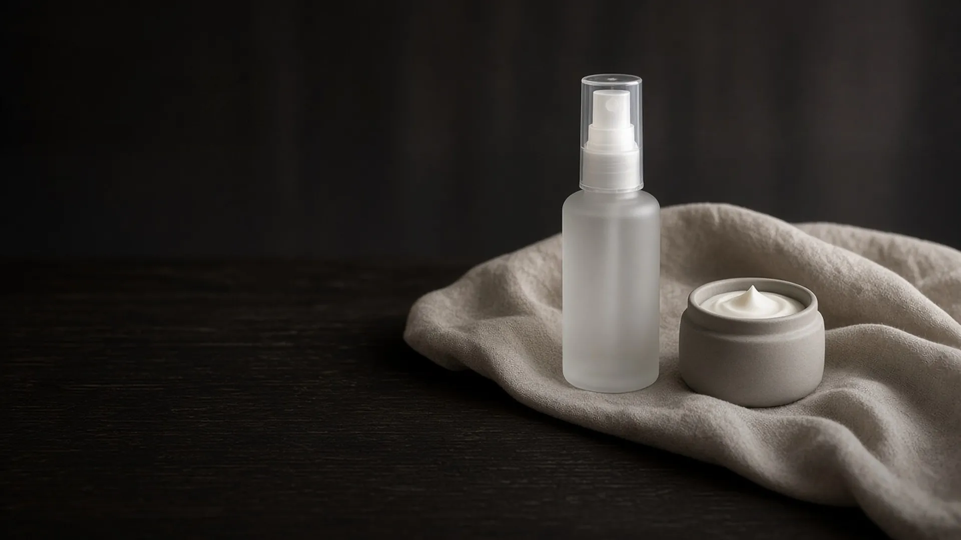 A plain frosted spray bottle beside a small open jar of white cream on linen